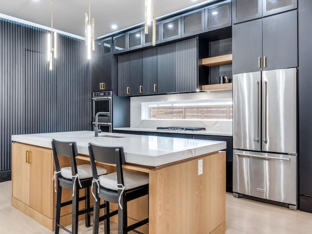 kitchen featuring light stone counters, stainless steel appliances, a sink, an island with sink, and pendant lighting