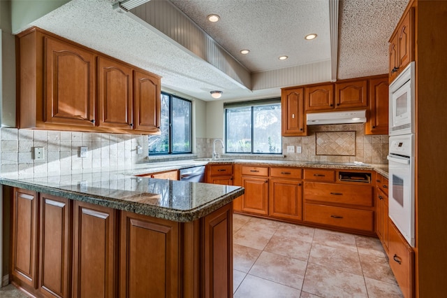 kitchen featuring a peninsula, white appliances, under cabinet range hood, and brown cabinets