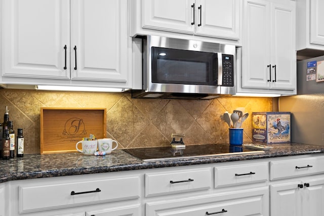 kitchen with black electric stovetop, stainless steel microwave, backsplash, white cabinetry, and dark stone countertops