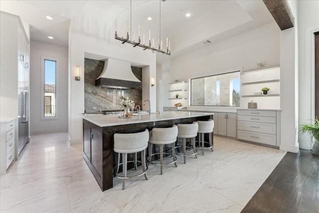 kitchen featuring premium range hood, a center island with sink, light countertops, and decorative backsplash