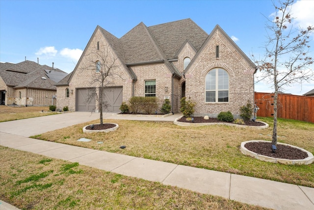 french provincial home featuring brick siding, concrete driveway, roof with shingles, fence, and a front yard