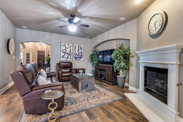 living room featuring a fireplace with raised hearth, baseboards, wood finished floors, arched walkways, and a ceiling fan