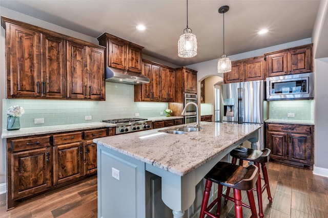 kitchen with light stone countertops, arched walkways, a sink, under cabinet range hood, and appliances with stainless steel finishes