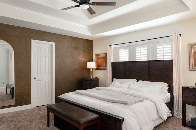 carpeted bedroom featuring arched walkways, a ceiling fan, baseboards, a raised ceiling, and crown molding