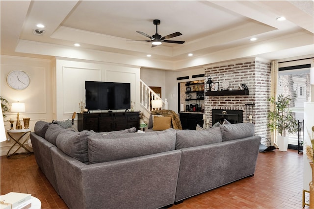 living area with a brick fireplace, visible vents, a raised ceiling, and crown molding