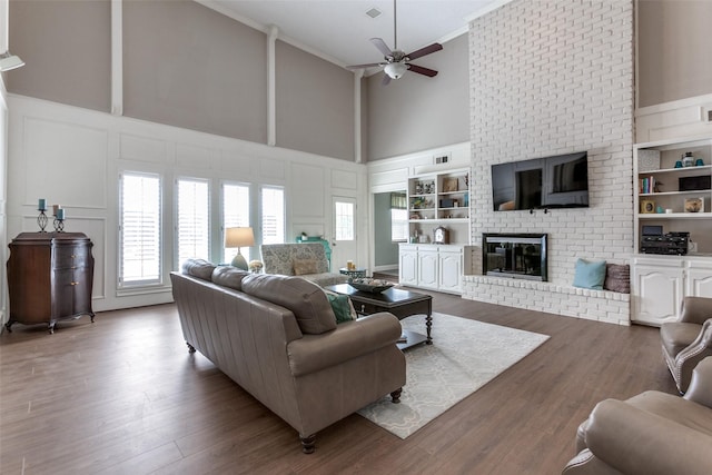 living room with a decorative wall, a towering ceiling, ceiling fan, a brick fireplace, and crown molding