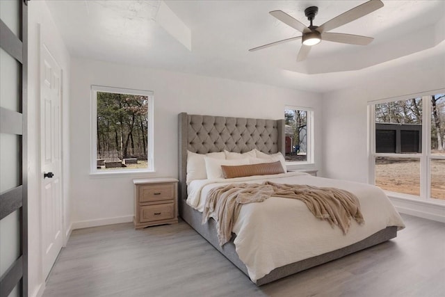 bedroom with light wood finished floors, baseboards, multiple windows, and a tray ceiling