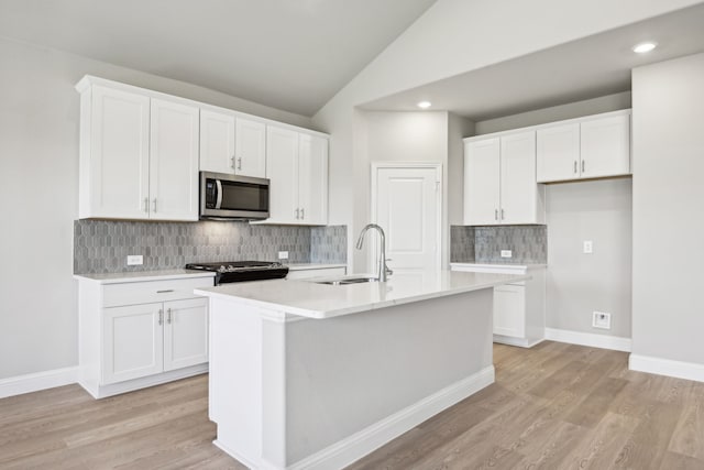 kitchen with a sink, stainless steel microwave, white cabinetry, vaulted ceiling, and an island with sink