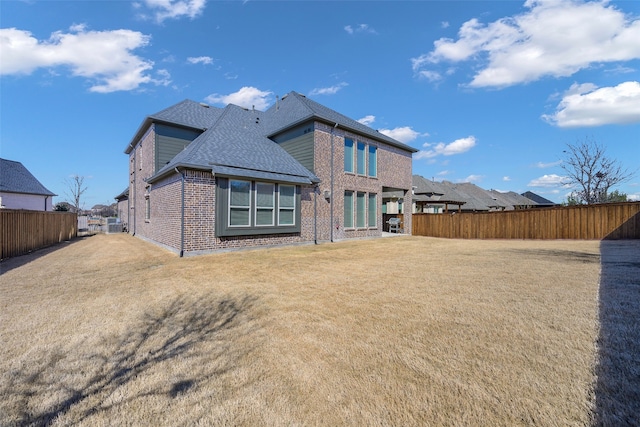 back of house featuring a shingled roof, fence, a lawn, and brick siding