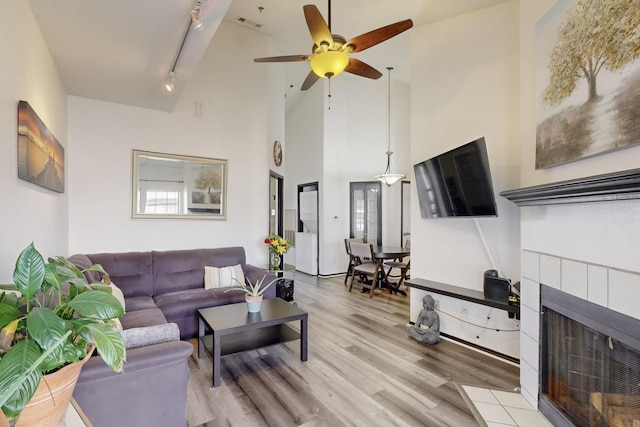 living room featuring track lighting, ceiling fan, light wood-style flooring, a tile fireplace, and a high ceiling