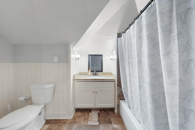 bathroom with shower / tub combo with curtain, toilet, stone finish floor, wainscoting, and vanity