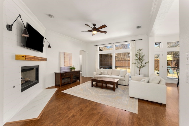 living area with arched walkways, a fireplace with flush hearth, wood finished floors, a ceiling fan, and ornamental molding