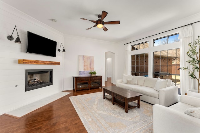 living room with arched walkways, a fireplace, wood finished floors, a ceiling fan, and ornamental molding