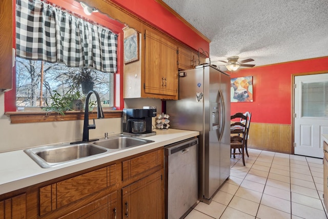 kitchen featuring a textured ceiling, a wainscoted wall, a sink, dishwasher, and brown cabinetry