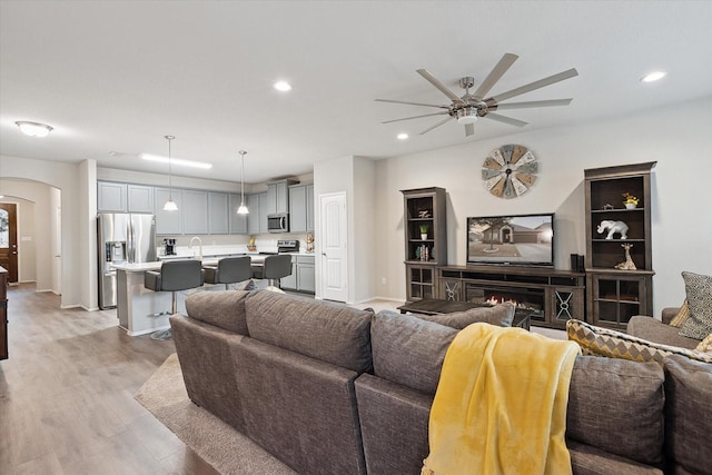 living area featuring arched walkways, recessed lighting, light wood-style flooring, a ceiling fan, and a lit fireplace