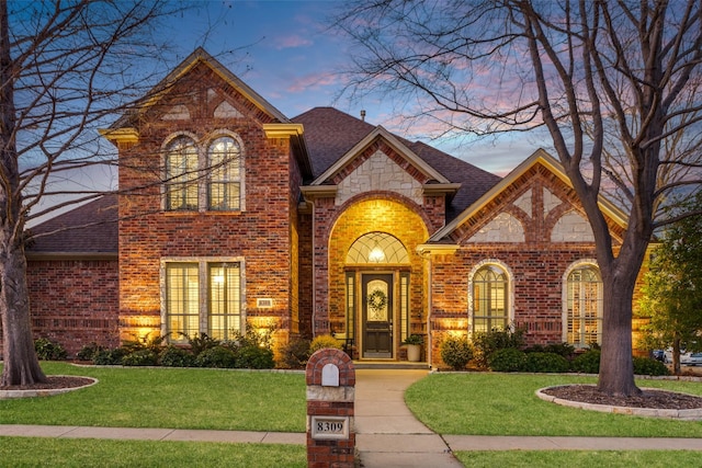 view of front of property with french doors, brick siding, roof with shingles, a lawn, and stone siding