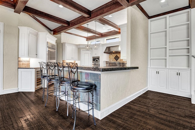 kitchen with white cabinetry, baseboards, wall chimney range hood, beam ceiling, and a kitchen bar