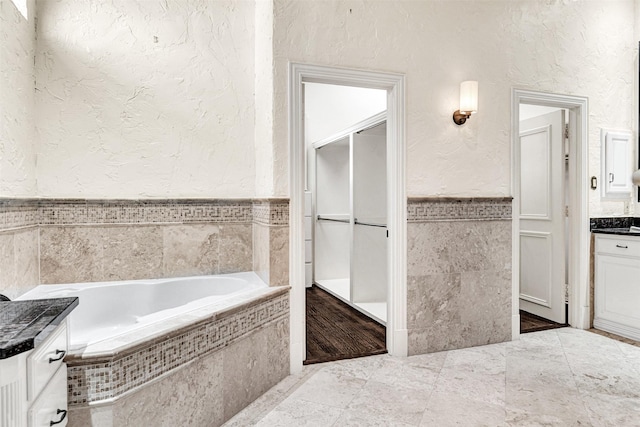 bathroom with a textured wall, a wainscoted wall, a garden tub, vanity, and tile walls