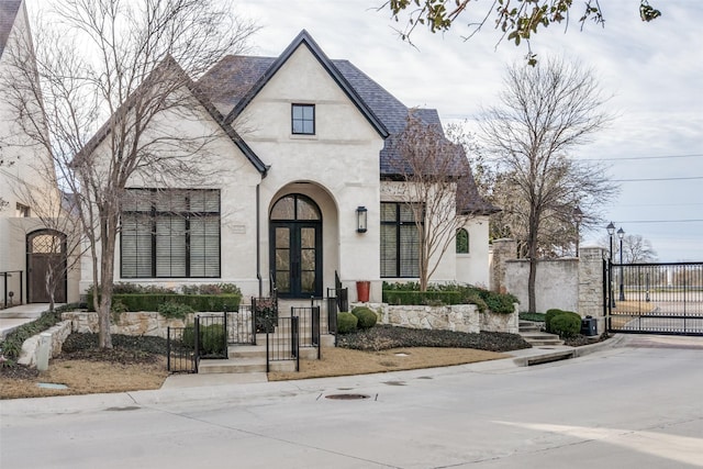 french country home with a shingled roof, a gate, french doors, and stucco siding