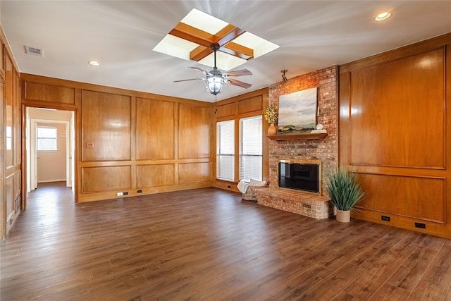 unfurnished living room with dark wood-style flooring, a skylight, a ceiling fan, visible vents, and a brick fireplace