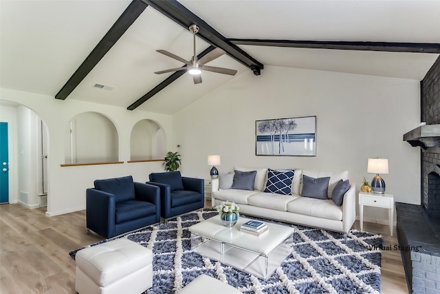 living area featuring arched walkways, ceiling fan, vaulted ceiling with beams, wood finished floors, and a brick fireplace