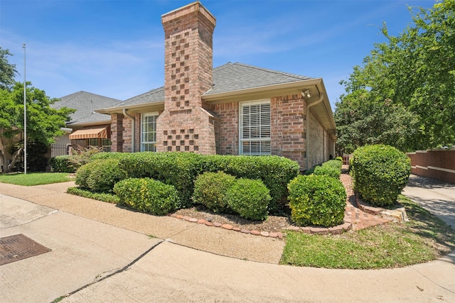 view of side of property with roof with shingles, brick siding, a chimney, and fence