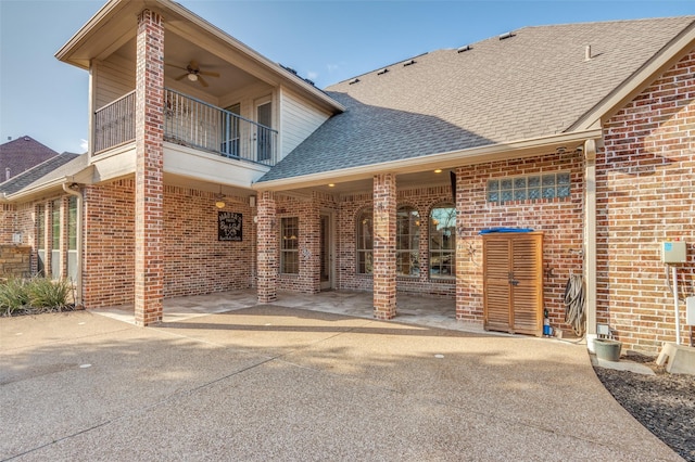 view of exterior entry featuring ceiling fan, a balcony, brick siding, a shingled roof, and a patio area