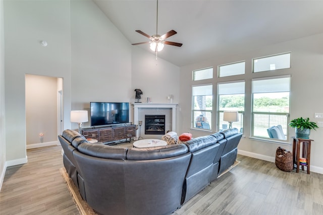 living room featuring baseboards, ceiling fan, light wood-style floors, a fireplace, and high vaulted ceiling