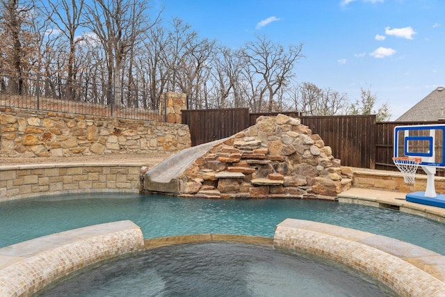 view of swimming pool with a water slide, fence, and a pool with connected hot tub