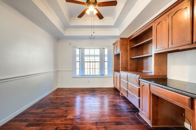 unfurnished office with a tray ceiling, dark wood-type flooring, ornamental molding, a ceiling fan, and baseboards