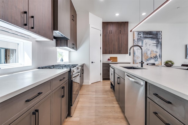 kitchen featuring a sink, light wood-style floors, light countertops, appliances with stainless steel finishes, and wall chimney exhaust hood