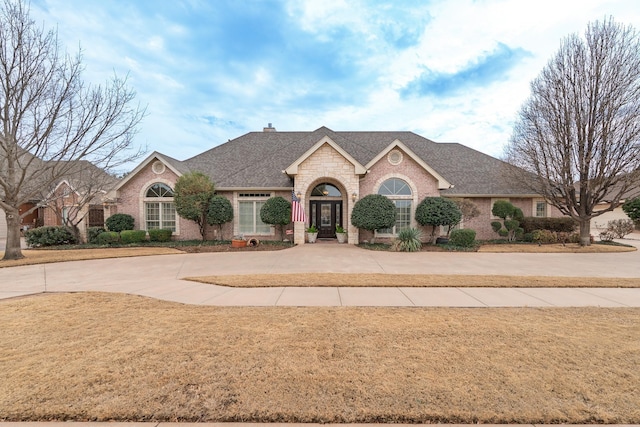 french country inspired facade featuring roof with shingles, a chimney, a front lawn, and brick siding