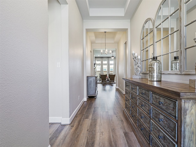 hall featuring dark wood-style floors, a tray ceiling, baseboards, and a notable chandelier