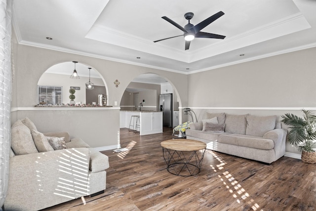 living room with dark wood-style floors, arched walkways, a raised ceiling, and a ceiling fan