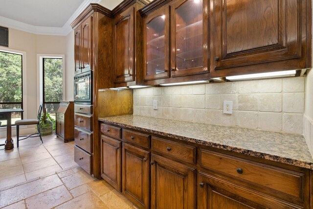 kitchen featuring stone countertops, backsplash, glass insert cabinets, crown molding, and stone tile flooring