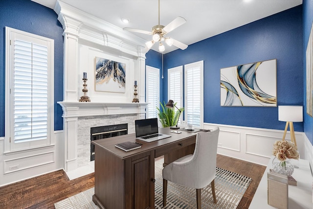 home office featuring dark wood-style floors, a wainscoted wall, a fireplace, and a ceiling fan