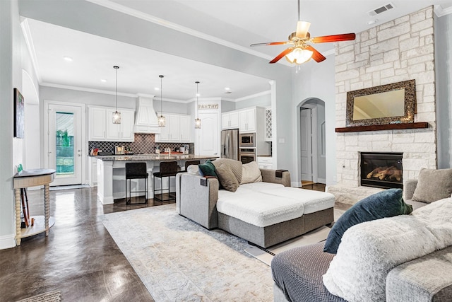 living room featuring baseboards, visible vents, arched walkways, crown molding, and a stone fireplace