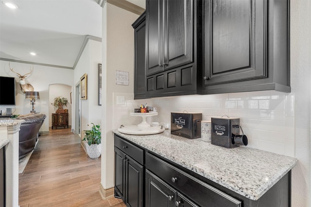 kitchen featuring arched walkways, dark cabinets, light wood-style floors, ornamental molding, and backsplash