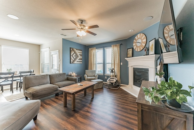 living area with dark wood-style flooring, visible vents, a glass covered fireplace, ceiling fan, and baseboards