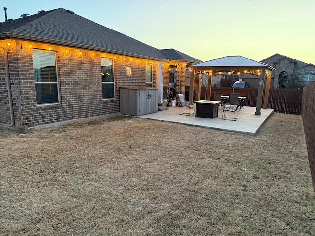 back of property at dusk with a fire pit, a fenced backyard, a gazebo, a patio area, and brick siding