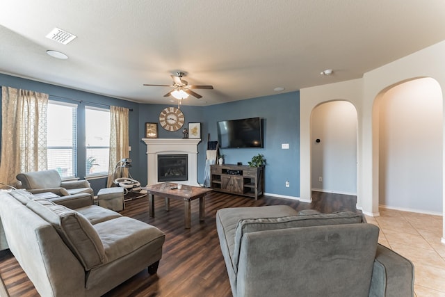 tiled living room featuring arched walkways, ceiling fan, visible vents, baseboards, and a glass covered fireplace