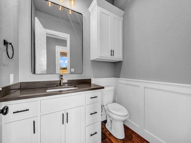 half bath featuring toilet, a decorative wall, wood finished floors, vanity, and wainscoting