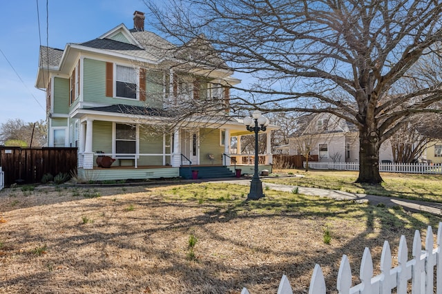 victorian home with a shingled roof, a chimney, fence private yard, covered porch, and a front lawn