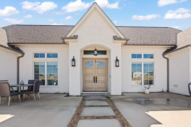 doorway to property featuring roof with shingles, brick siding, and a patio