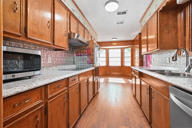 kitchen featuring stainless steel appliances, a sink, under cabinet range hood, a textured ceiling, and brown cabinets