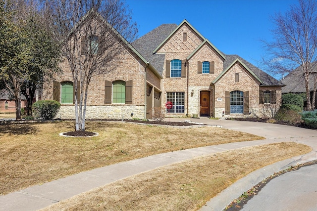 french provincial home featuring brick siding, a shingled roof, concrete driveway, stone siding, and a front yard