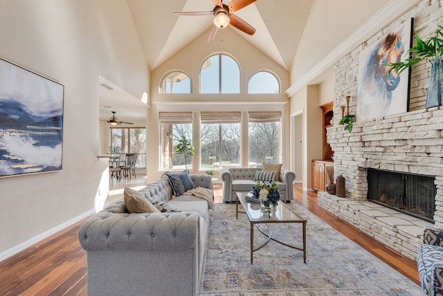 living area featuring a fireplace, a ceiling fan, wood finished floors, high vaulted ceiling, and baseboards