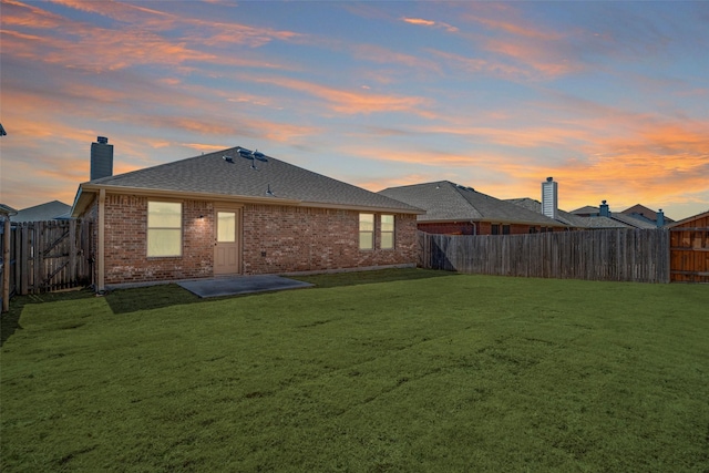back of property at dusk featuring a lawn, a chimney, brick siding, and a fenced backyard