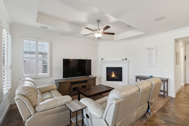 living room with a ceiling fan, baseboards, ornamental molding, a raised ceiling, and a glass covered fireplace