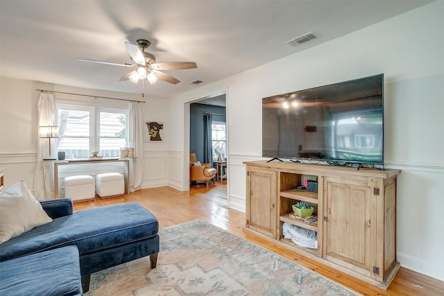 living area featuring light wood-type flooring, visible vents, a ceiling fan, and wainscoting